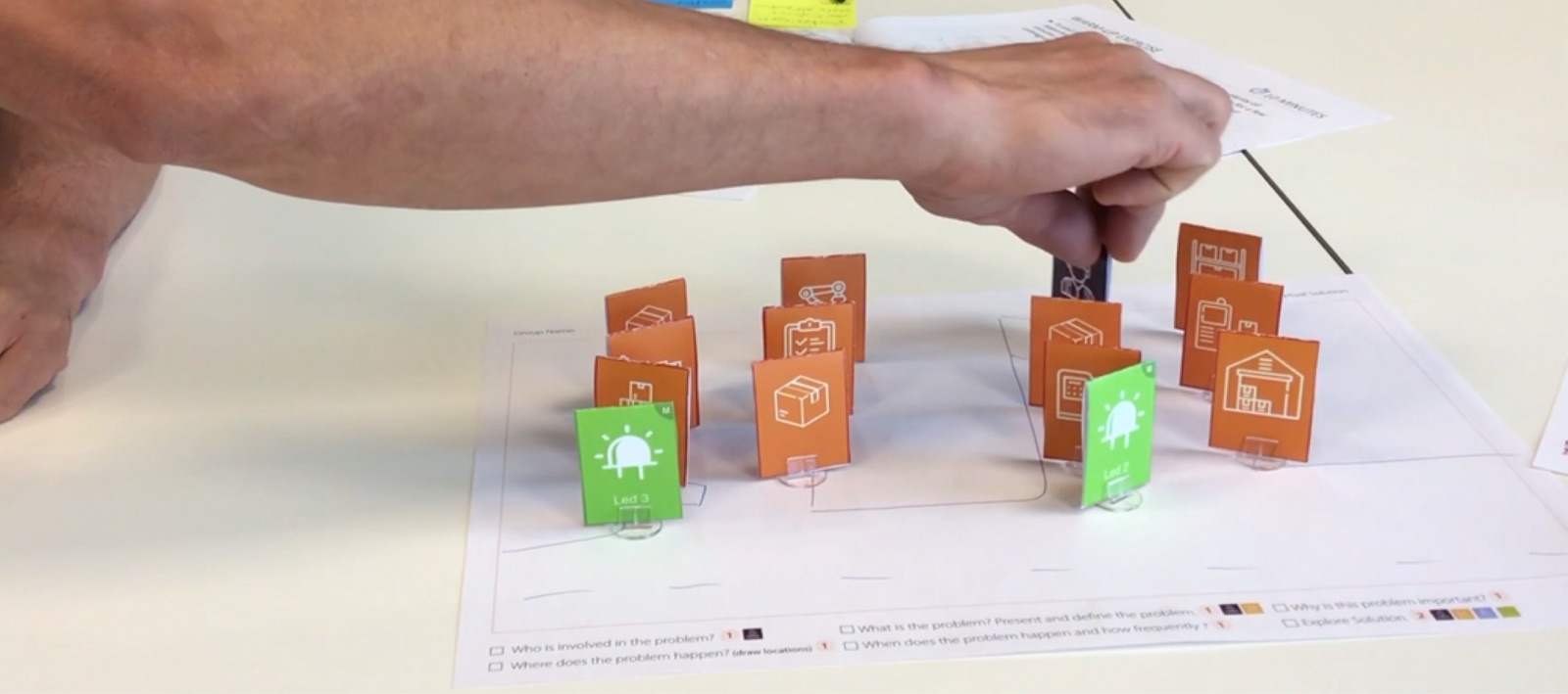 An arm reaches over a canvas full of orange tokens to move a dark purple worker token between two green lamp tokens