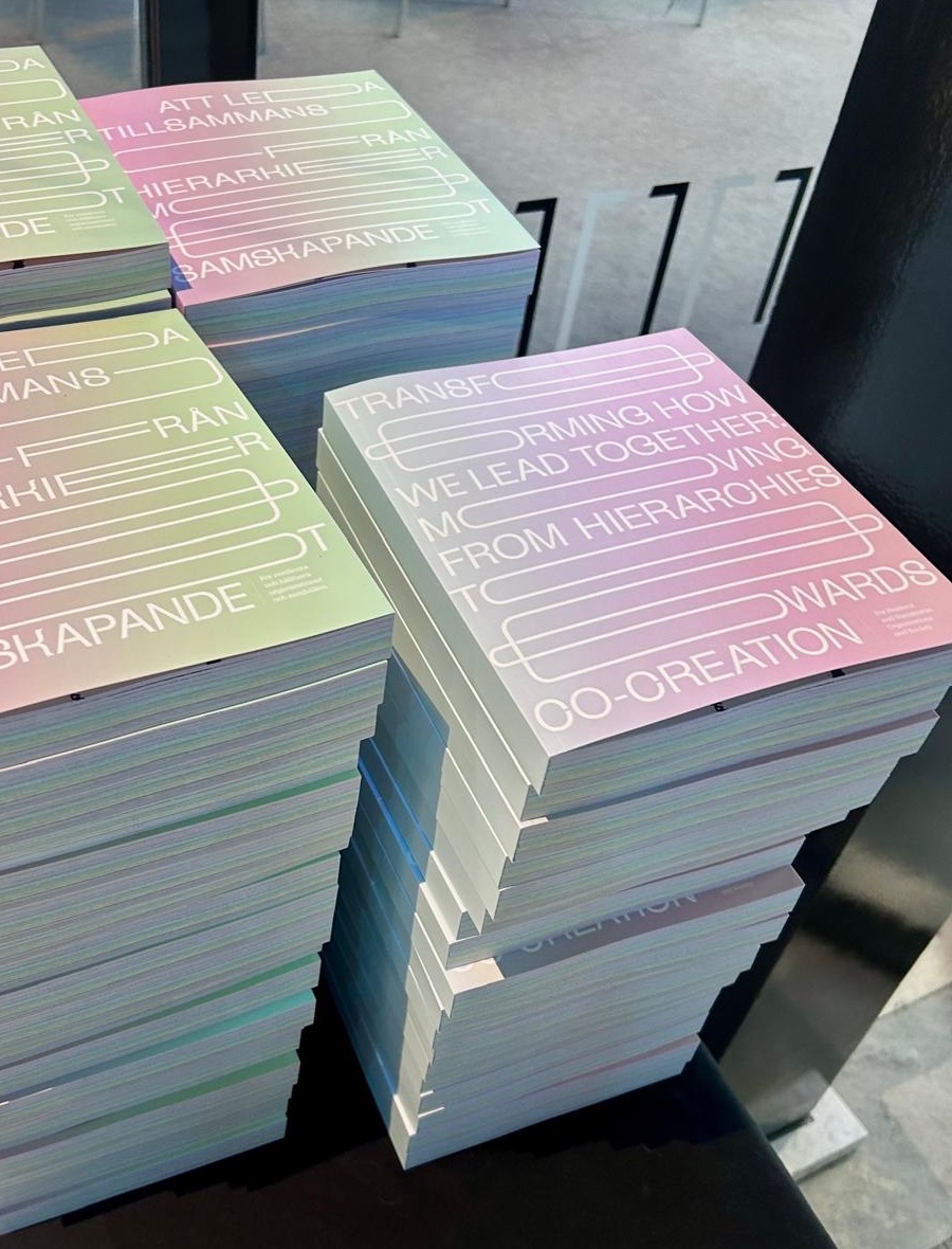 Stacks of the handbook on a table: the Swedish edition, Att leda tillsammans, in green and pink, and the English edition, Transforming how we lead together, in pink and lilac